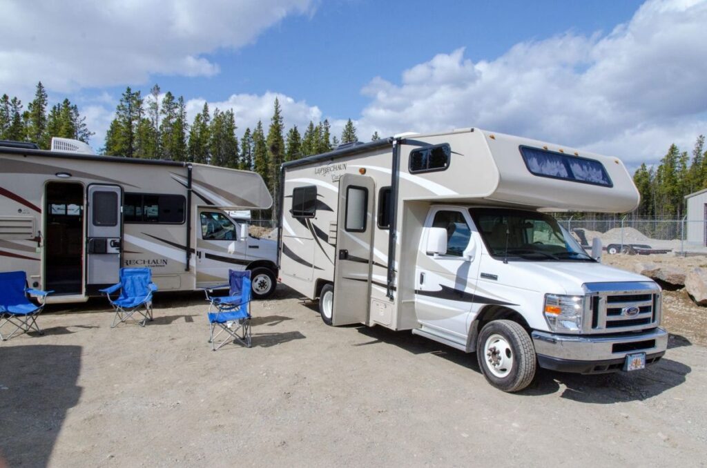 Two motorhomes with four camping chairs in front standing on a gravel parking lot with trees in the background