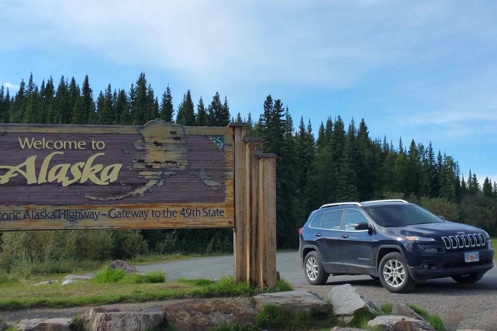 lateral view of a blue SUV during daylight on the right side of a welcome to alaska sign and in front of trees
