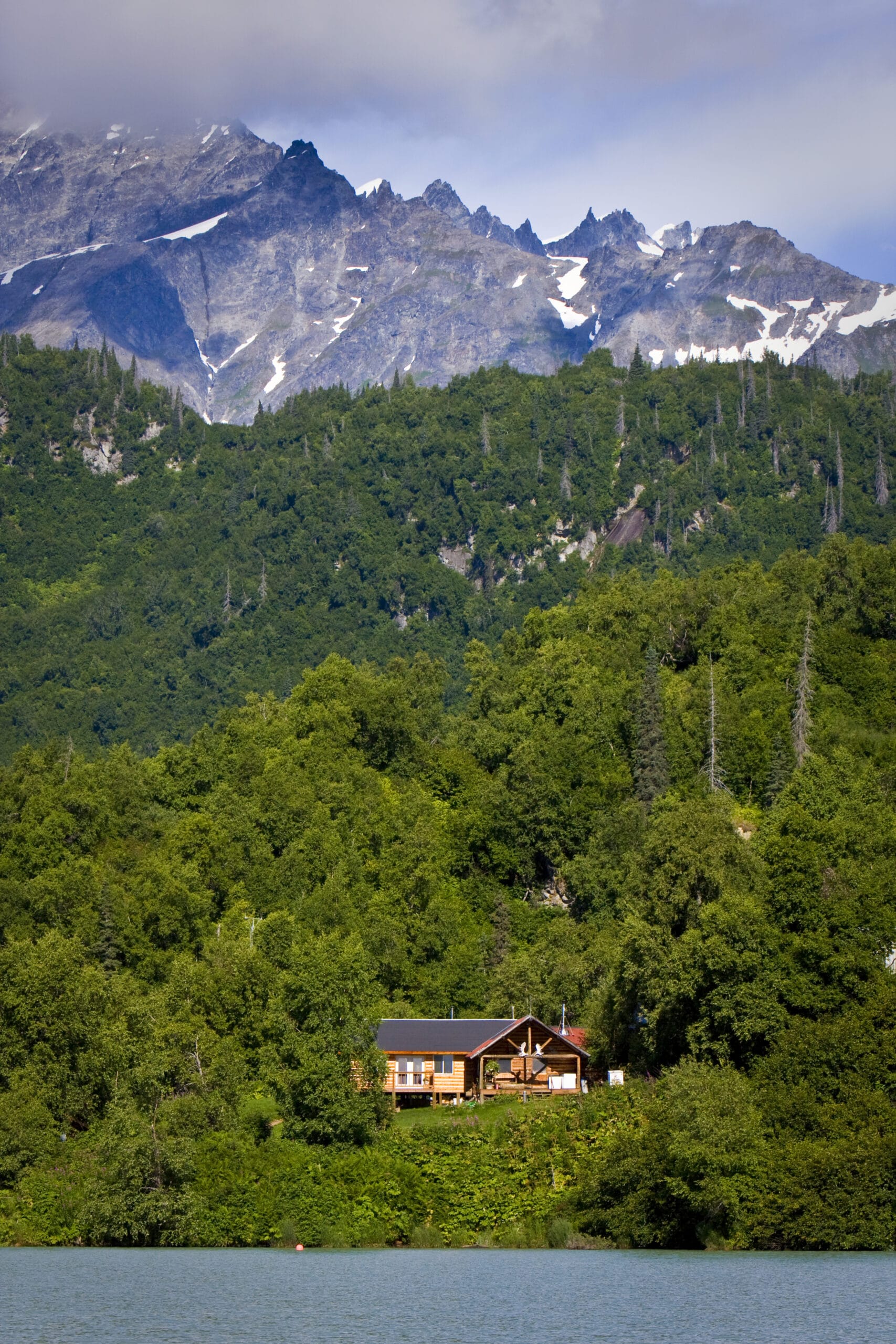 landscape in the front the see, in the middle a lodge among trees and in the background mountains with blue sky