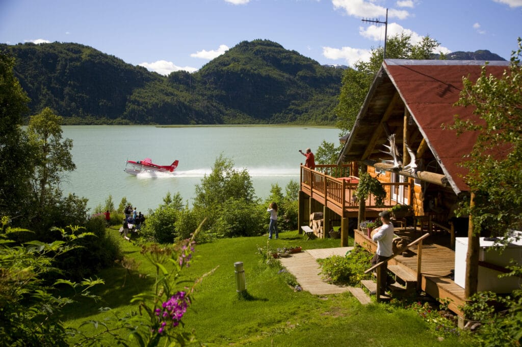 A lodge on the right with a lwan a seaplane starting on the sea and mountains in the background