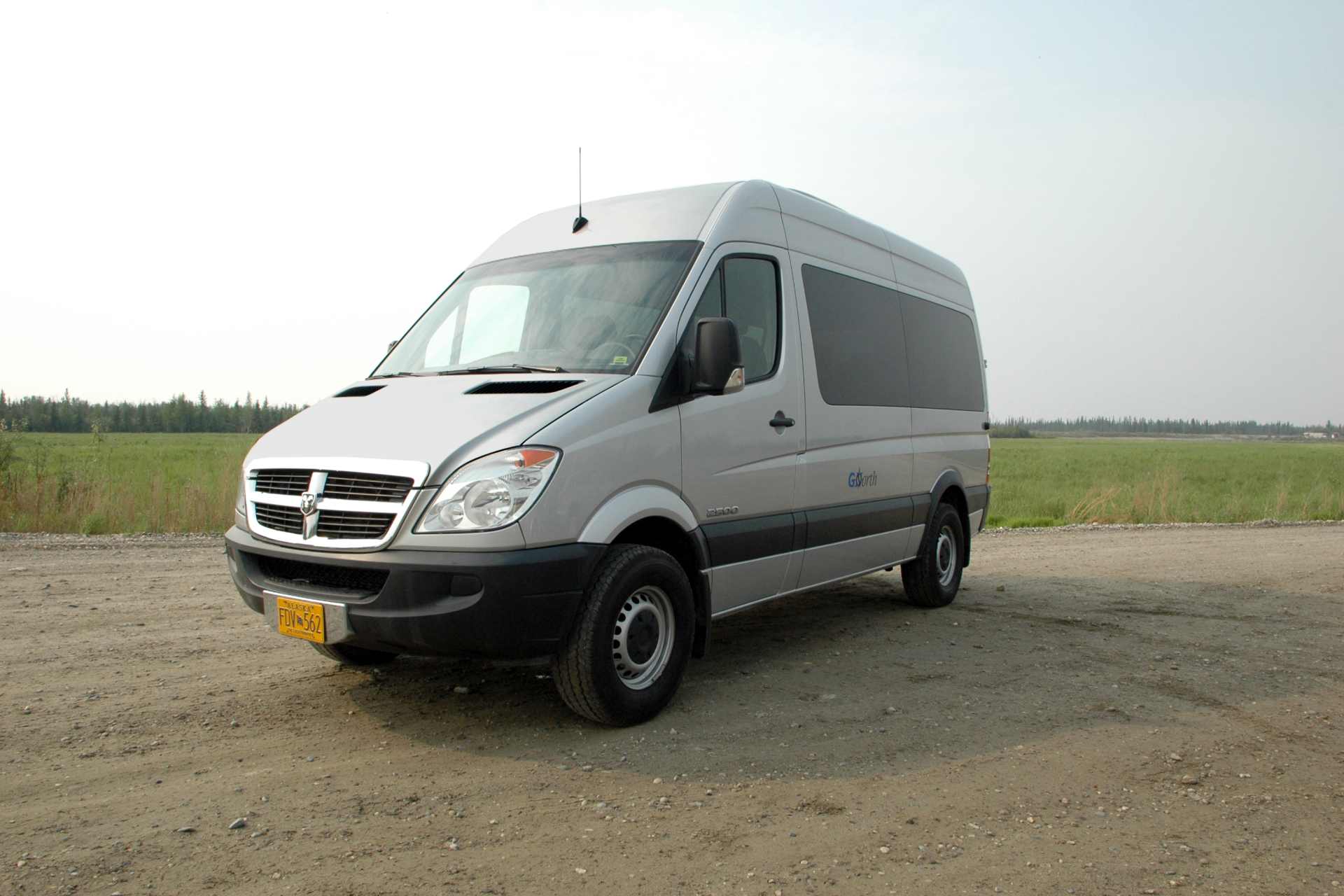 lateral view of a grey passenger van during daylight on a gravel road and a green plain in the background