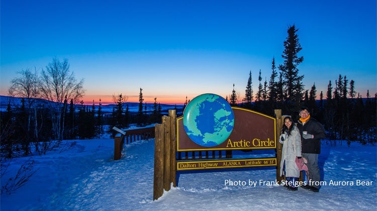 2 people standing at the Arctic Circle sign in snow with beautiful blue and orange sky