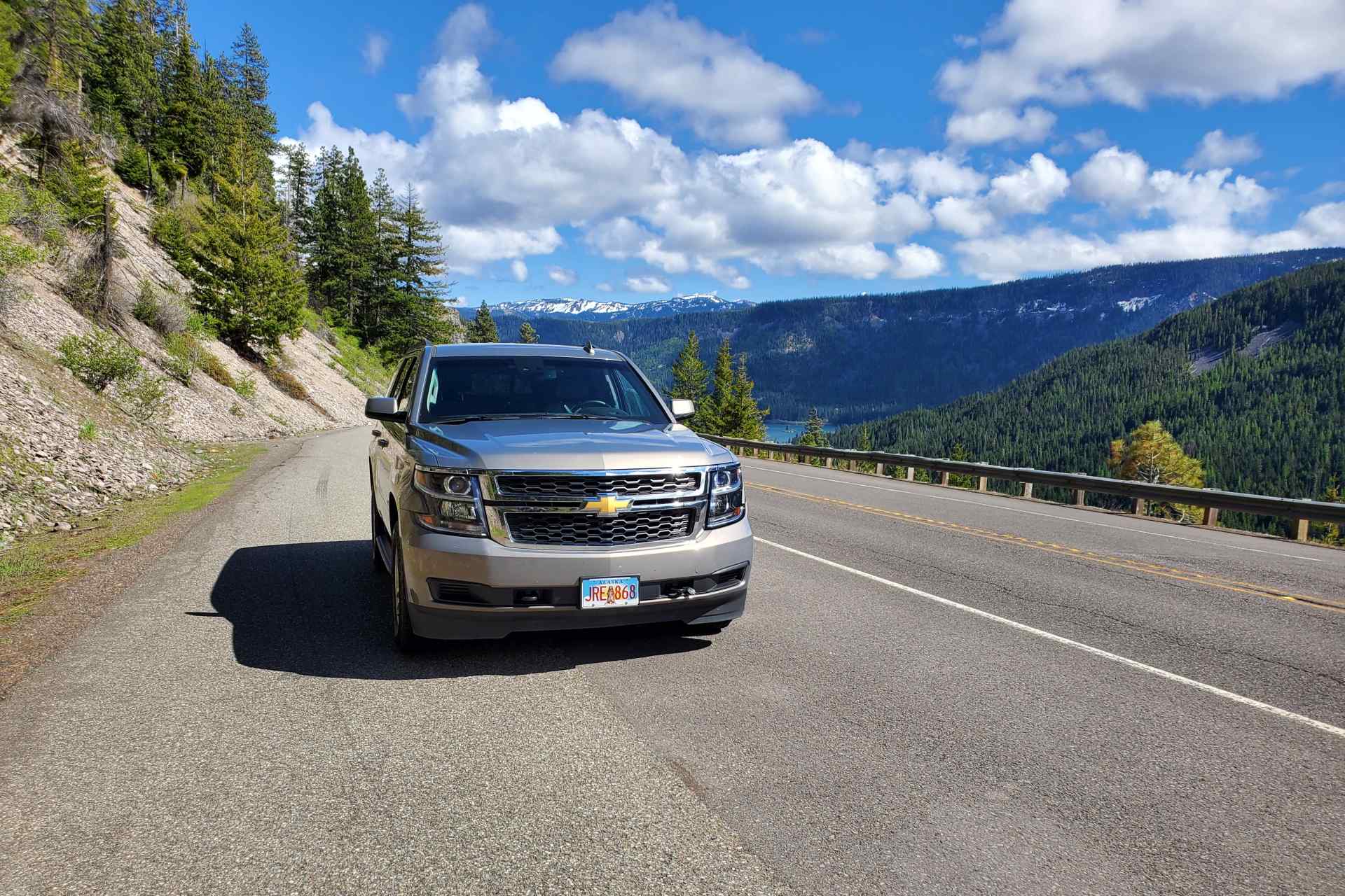 an SUV during daylight on the side of a road with trees on the hillside on the left and green hills in the background