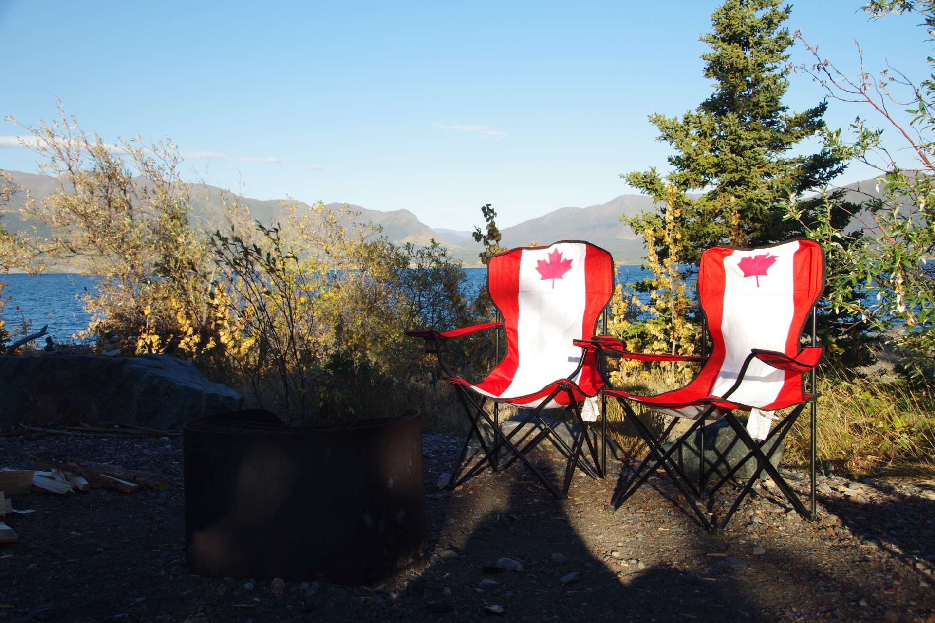 Camping chairs during daylight in the center of the picture with the canadian flag printed on them and with a lake, some trees and a hill range in the background