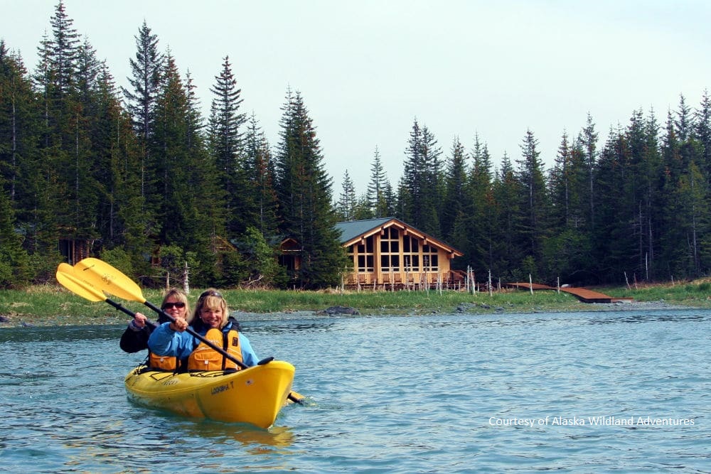 2 people in a yellow kayak on the sea and a lodge surrounded by trees in the back