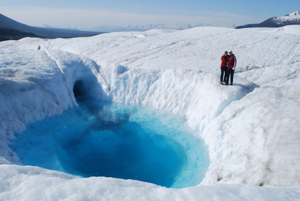 two people standing at a blue whole on a glacier