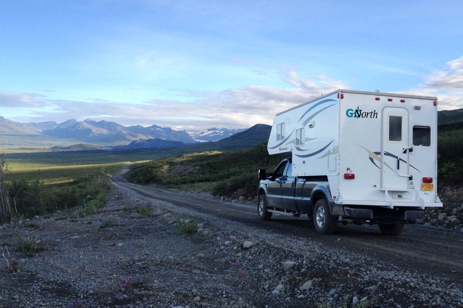 Back view of a truck camper on a gravel road with a green plain on the left and a mountain range in the background