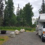 A motorhome on the right side parked in a gravel lot next to a fire pit surrounded by trees during daylight