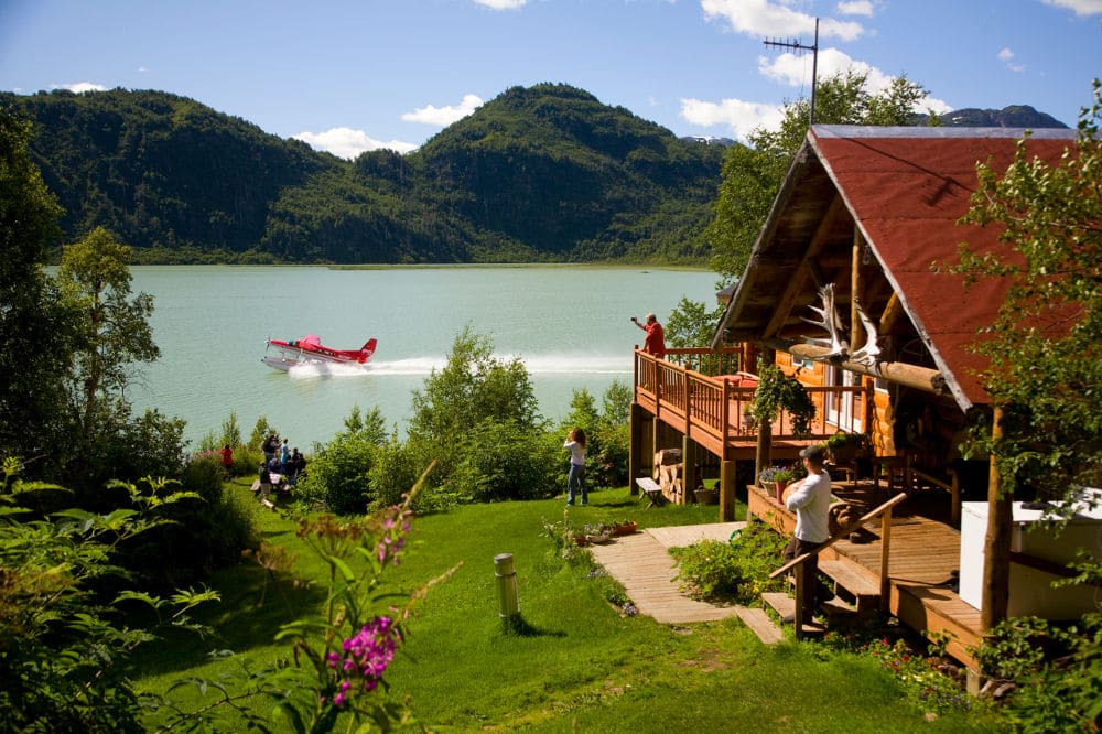 lodge in beautiful setting with mountains in the background, a seaplane starting in the middle and a ncier garden in the front