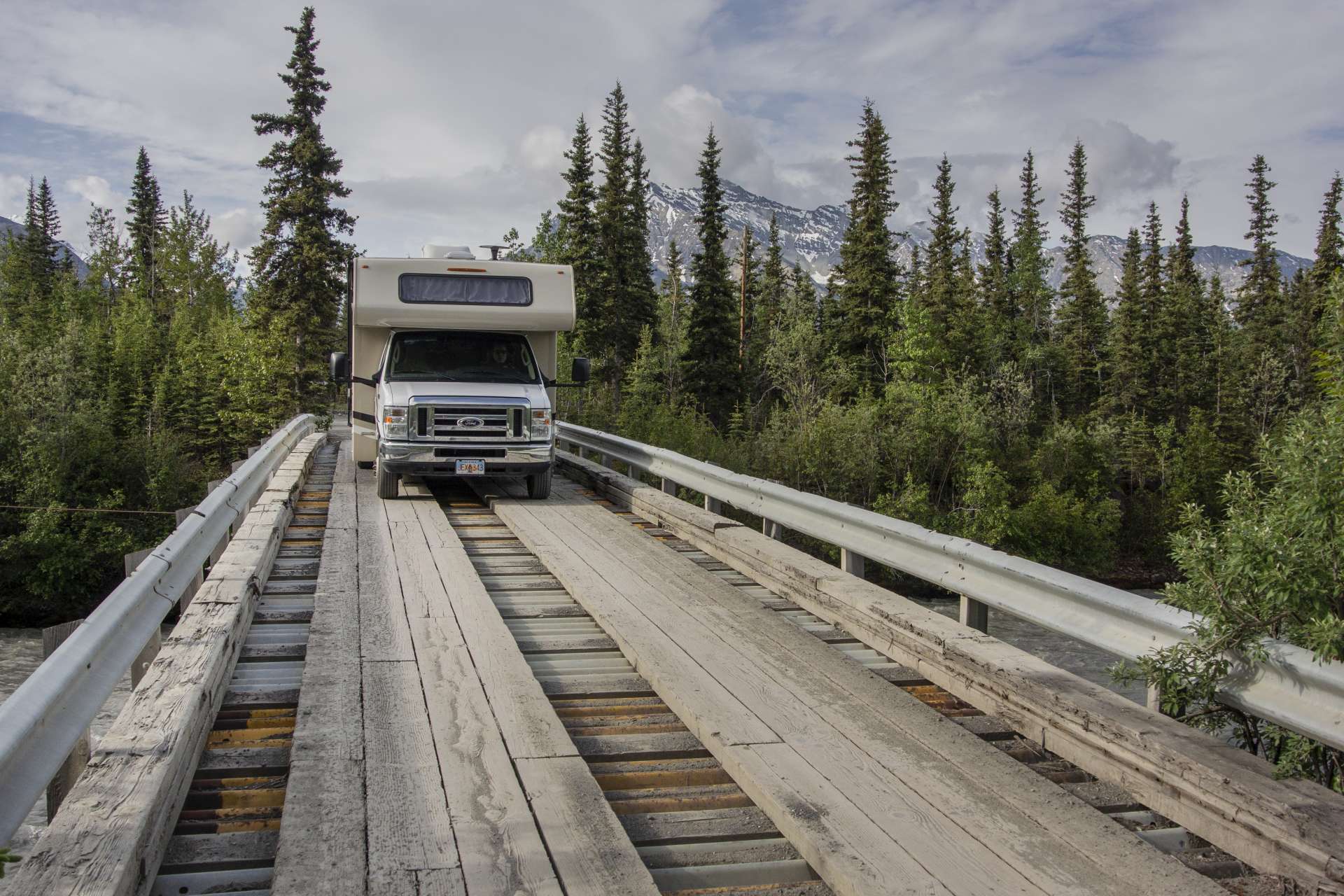 A motorhome driving on a wooden bridge with trees and a mountain in the background during daylight